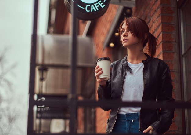 Una hermosa chica con una chaqueta de cuero con una mochila sosteniendo una taza con café para llevar afuera cerca del café.