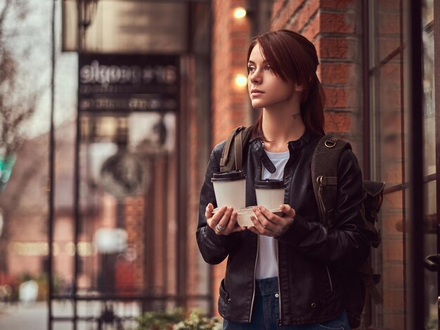 Una hermosa chica con una chaqueta de cuero con una mochila sosteniendo tazas con café para llevar afuera cerca del café.