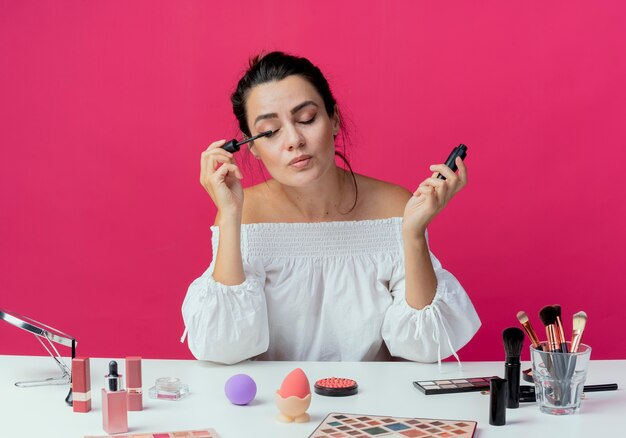 Hermosa chica complacida se sienta a la mesa con herramientas de maquillaje aplicando rímel con los ojos cerrados aislados en la pared rosa