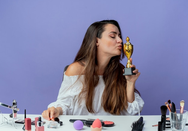 Hermosa chica complacida se sienta a la mesa con herramientas de maquillaje finge besar la copa ganadora aislada en la pared púrpura