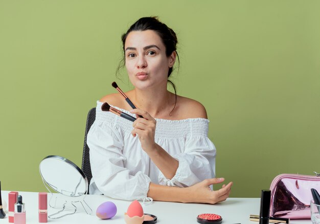 Hermosa chica complacida se sienta a la mesa con herramientas de maquillaje sosteniendo pinceles de maquillaje aislados en la pared verde