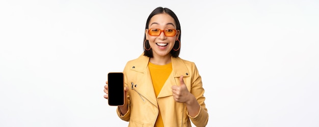 Hermosa chica coreana mujer asiática en gafas de sol que muestra la interfaz de la aplicación del teléfono inteligente pulgares arriba recomendando la aplicación del teléfono móvil fondo blanco
