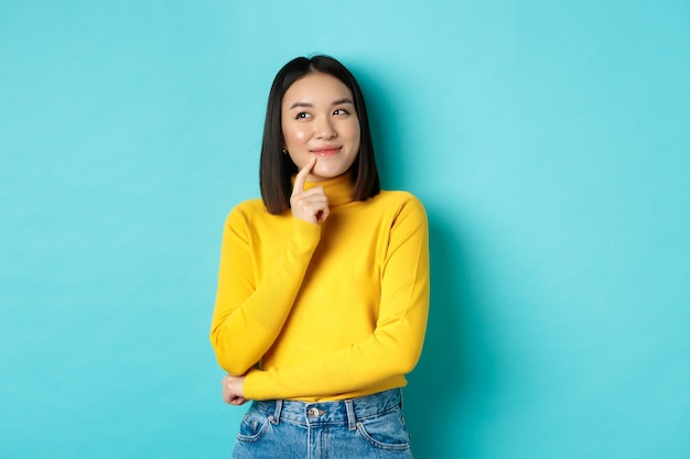 Hermosa chica coreana pensando, imaginando y sonriendo, mirando la esquina superior izquierda y soñando despierto, de pie contra el fondo azul