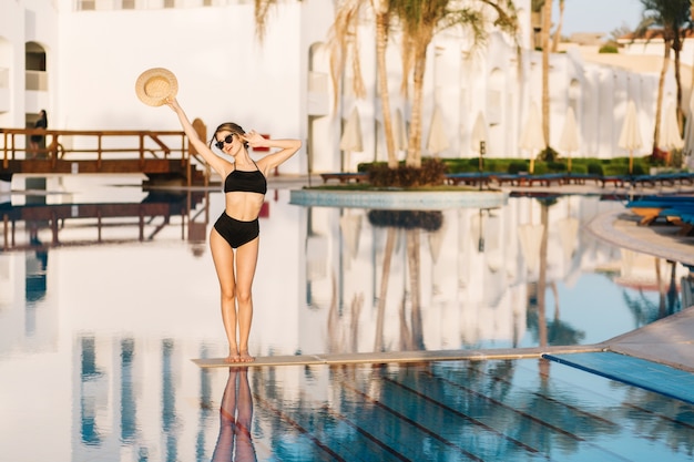 Hermosa chica con cuerpo delgado, modelo con traje de baño negro posando en medio de la piscina en hotel de lujo, resort. Vacaciones, vacaciones, verano.