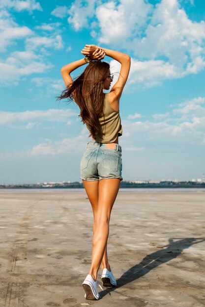 Hermosa chica delgada fitness bronceado está de vuelta con las manos en la parte superior. Posando al aire libre en pantalones vaqueros con estilo. Luz tenue.