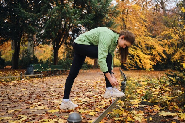 Hermosa chica deportiva atando cordones de zapatos en zapatillas antes de correr en el acogedor parque de otoño