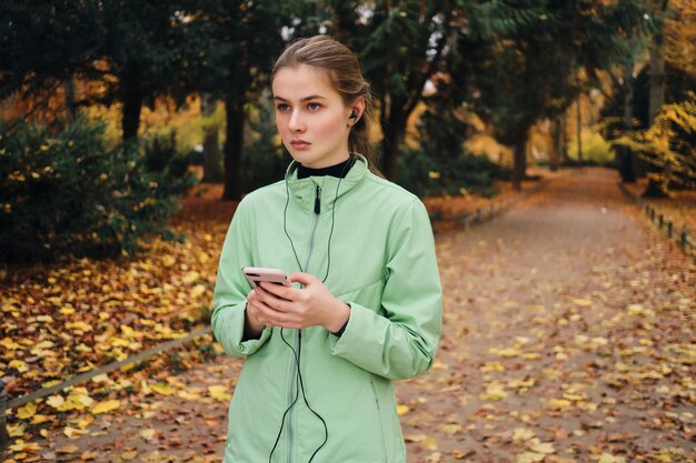 Hermosa chica deportiva en auriculares con celular descansando después de correr en el acogedor parque de otoño