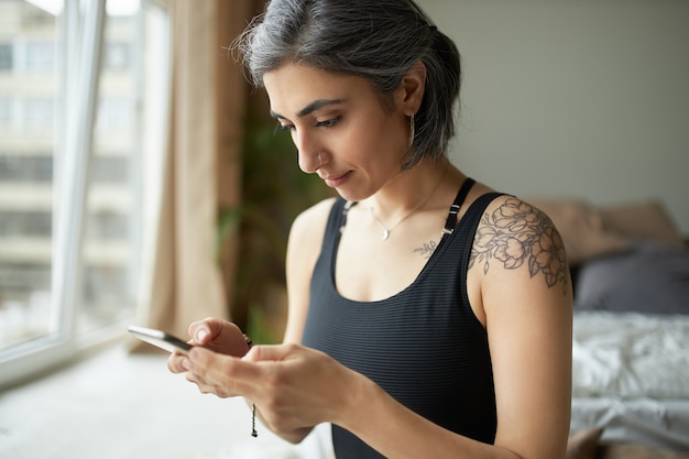 Hermosa chica deportiva con cabello gris prematuro, tatuaje en el hombro y piercing en la nariz sentado en el interior con teléfono móvil