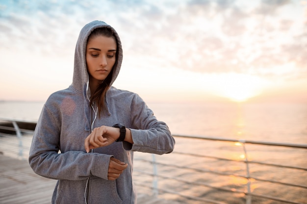 Hermosa chica deportiva mirando el reloj durante el amanecer sobre la playa.