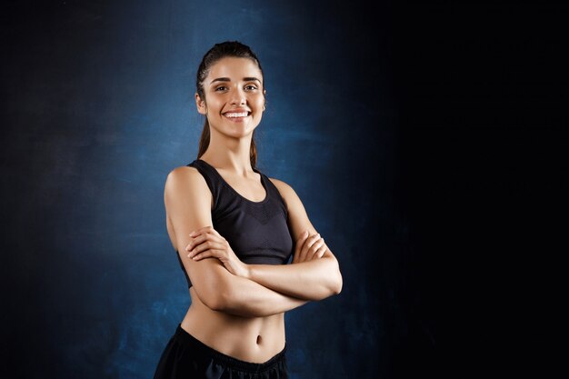 Hermosa chica deportiva posando con los brazos cruzados sobre la pared oscura.