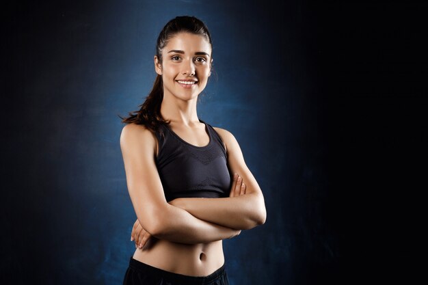 Hermosa chica deportiva posando con los brazos cruzados sobre la pared oscura.