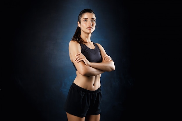 Hermosa chica deportiva posando con los brazos cruzados sobre la pared oscura.