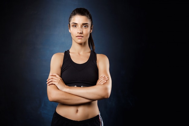 Hermosa chica deportiva posando con los brazos cruzados sobre la pared oscura.