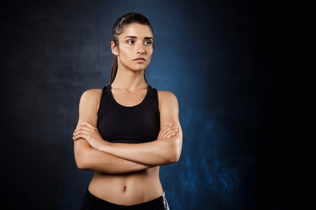 Hermosa chica deportiva posando con los brazos cruzados sobre la pared oscura.