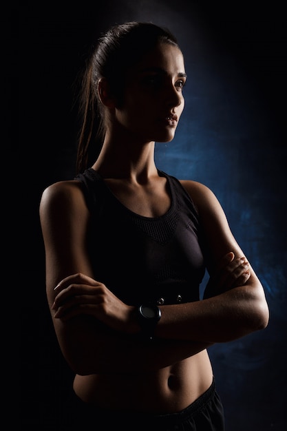 Hermosa chica deportiva posando con los brazos cruzados sobre la pared oscura.