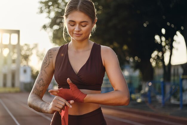 Hermosa chica deportiva en ropa deportiva envolviendo cuidadosamente el vendaje de boxeo rojo a mano en el estadio de la ciudad