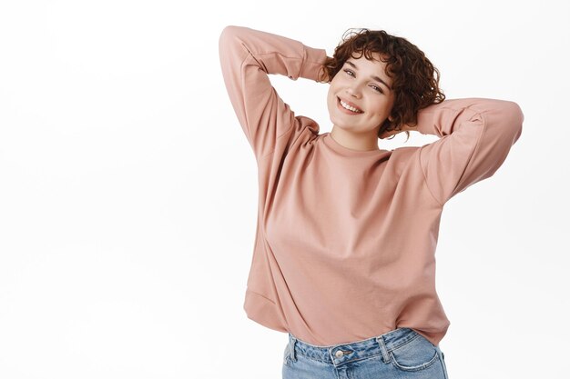 Hermosa chica descansando y relajada sonriendo, tomándose de la mano detrás de la cabeza y relajándose, descansando, disfrutando del tiempo libre, de pie contra el fondo blanco