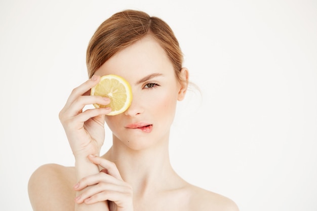 Hermosa chica desnuda con la piel limpia y sana mordiendo el labio ocultando el ojo detrás de la rodaja de limón. Spa de belleza cosmetología.