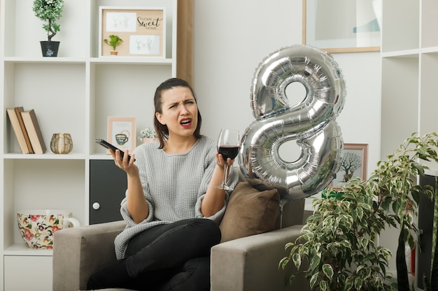 Foto gratuita hermosa chica disgustada en el día de la mujer feliz sosteniendo una copa de vino con el teléfono sentado en un sillón en la sala de estar