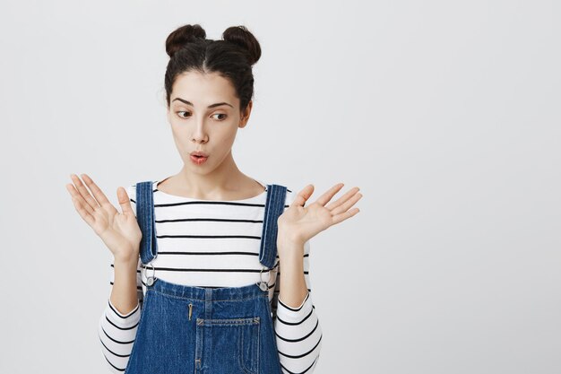 Hermosa chica con dos moños tiene una apariencia atractiva y atractiva y una expresión de asombro, haciendo pucheros con los labios de emoción mientras recibe noticias inesperadas.