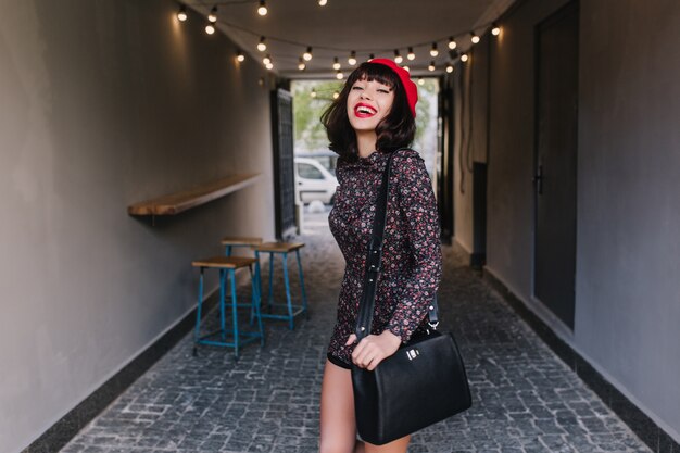 Hermosa chica elegante con cabello corto oscuro caminando después del trabajo con bolso de cuero negro y sonriendo. Retrato de adorable joven morena vistiendo ropa francesa vintage, divirtiéndose en el interior