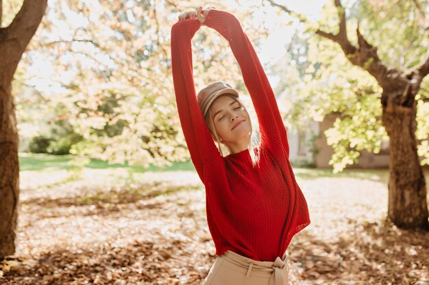 Hermosa chica elegante sonriendo en el parque bajo el sol. Encantadora rubia en suéter rojo sintiéndose feliz al aire libre.