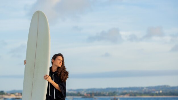 Hermosa chica se encuentra en la playa con una tabla de surf.