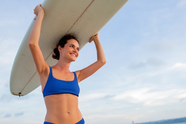 Hermosa chica se encuentra en la playa con una tabla de surf.