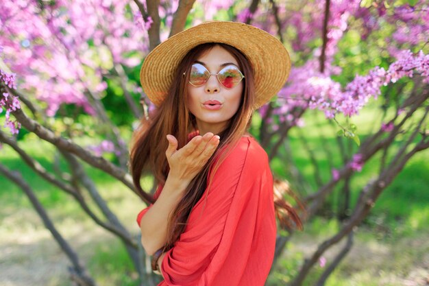 Hermosa chica enviando beso de aire a la cámara en un día soleado de primavera. Árboles en flor rosa o