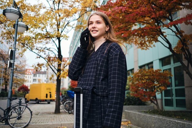 Hermosa chica con estilo felizmente hablando por teléfono celular mientras está de pie en la calle de la ciudad con la maleta