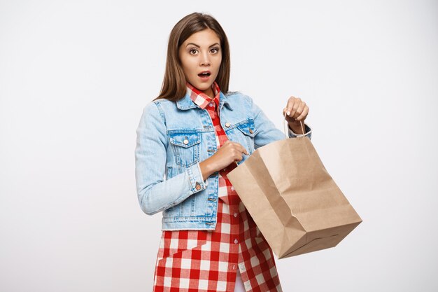 Hermosa chica con estilo que parece sorprendida después de abrir la bolsa de papel