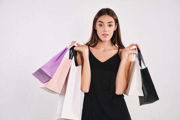 Hermosa chica con estilo en vestido negro sosteniendo bolsas de compras mirando atentamente en cámara sobre fondo blanco aislado
