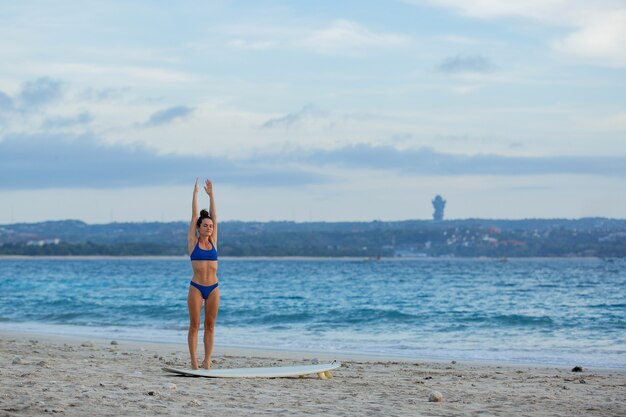 Hermosa chica estirándose en la playa con una tabla de surf.