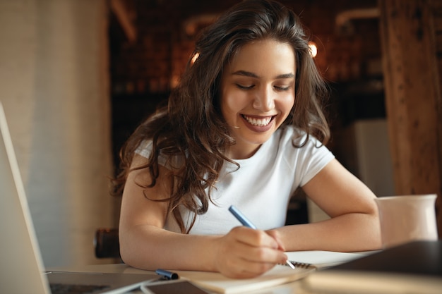 Foto gratuita hermosa chica estudiante con mejillas regordetas sosteniendo lápiz de escritura a mano en el cuaderno mientras aprende a distancia de casa