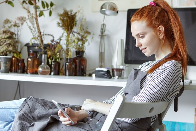 Hermosa chica estudiante pelirroja que participa en un retiro de pintura, estudia nuevas técnicas de arte, se sienta en una silla en el taller y toma notas en su dispositivo, habiendo inspirado la expresión de su rostro