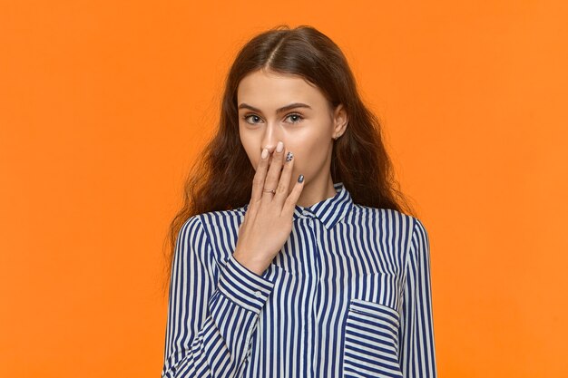 Hermosa chica europea esbelta con cabello oscuro y rizado posando en la pared naranja con la mano en los labios