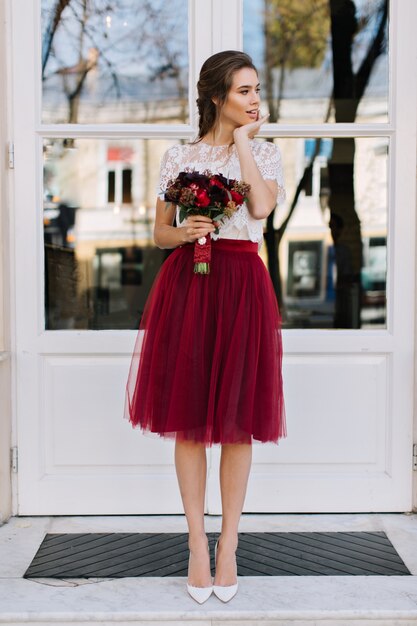 Hermosa chica en falda de tul marsala en tacones caminando en la calle. Ella sostiene boquet de flores y sonriendo a un lado