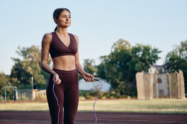 Hermosa chica fitness en ropa deportiva elegante con saltar la cuerda entrenando soñadoramente en el estadio de la ciudad