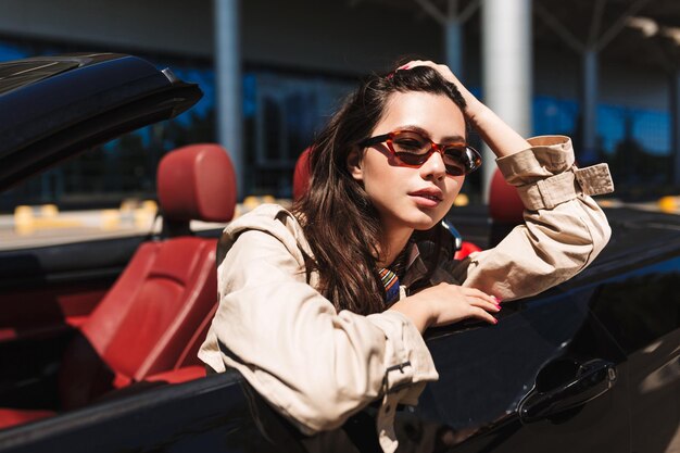 Hermosa chica con gafas de sol apoyada en la puerta del coche descapotable mientras mira soñadoramente a la cámara con el aeropuerto de fondo