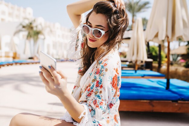 Hermosa chica en gafas de sol blancas de moda escribiendo un mensaje a un amigo, sosteniendo su cabello oscuro. Retrato de mujer joven morena increíble en camisa elegante sentado afuera con teléfono