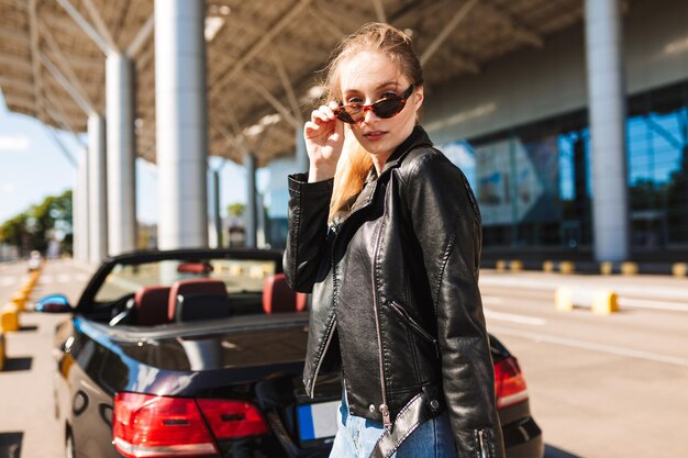 Hermosa chica con gafas de sol y chaqueta de cuero mirando cuidadosamente a la cámara cerca del aeropuerto con un auto descapotable negro en el fondo