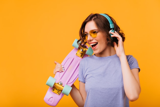 Hermosa chica con gafas de sol de color amarillo escuchando música en auriculares. Retrato de modelo femenino de moda con patineta cantando su canción favorita.