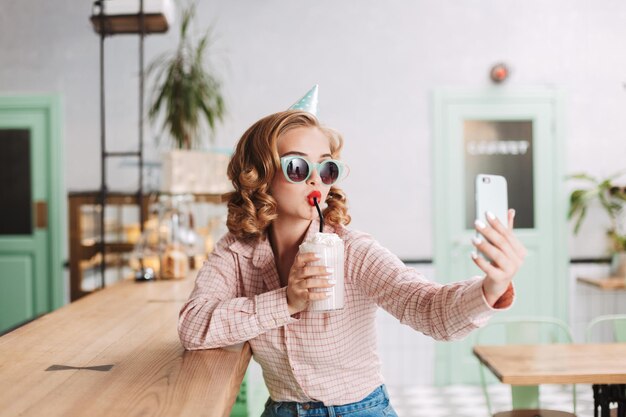 Hermosa chica con gafas de sol y gorra de cumpleaños sentada en el mostrador del bar y bebiendo batido mientras toma lindas fotos en su celular en el café