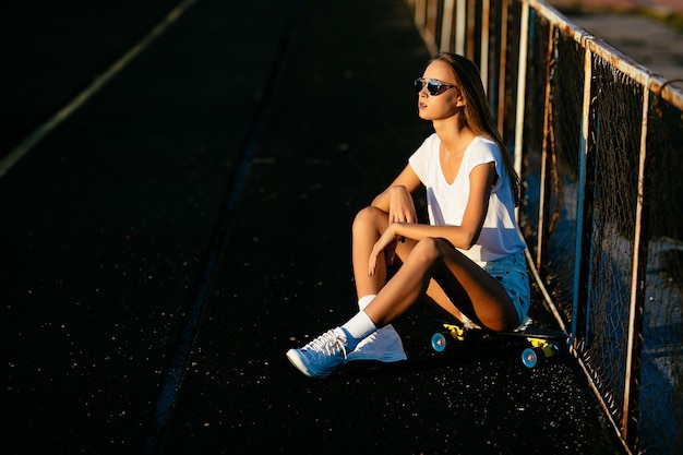 Foto gratuita una hermosa chica en las gafas de sol sentado cerca de la valla en su patineta en la puesta del sol.