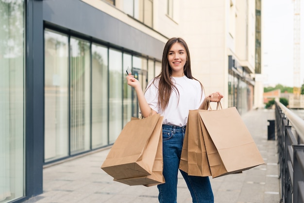 Hermosa chica en gafas de sol sostiene bolsas de la compra y sonríe mientras camina por la calle