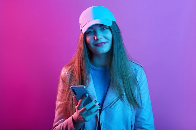 Hermosa chica con gorra de canasta y chaqueta de cuero, sosteniendo un moderno teléfono inteligente en las manos, mirando sonriendo directamente a la cámara