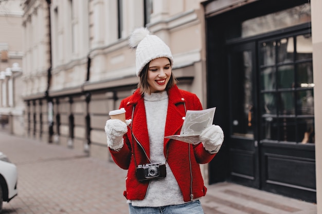 Hermosa chica con gorro de punto y guantes examina el mapa de lugares de interés. Mujer en abrigo rojo con vidrio de cartón y cámara retro.