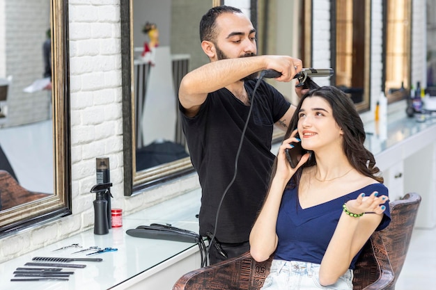 Hermosa chica hablando por teléfono mientras el peluquero le da forma al cabello