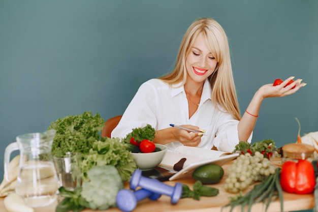 Hermosa chica hace una ensalada. Rubia deportiva en una cocina. Receta de escritura de mujer en el cuaderno.