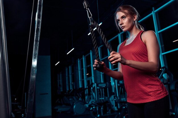 Hermosa chica haciendo ejercicios en el simulador en el gimnasio vestida con camiseta sin mangas rosa en blanco y medias. Sobre un fondo oscuro el gimnasio. Vestido con ropa deportiva.